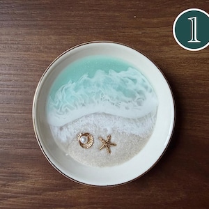Puede incluir: Un plato redondo de cerámica blanca con un diseño de resina que imita una escena de playa con agua turquesa, olas de espuma blanca y arena. Incluye una pequeña concha marina dorada con una perla blanca y una estrella de mar dorada.