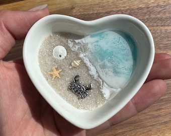 May include: A white heart-shaped dish with a blue and white resin ocean scene. The dish features a seashell, a starfish, and a silver crab.