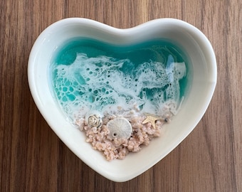 May include: A white heart-shaped dish with a blue and white resin ocean scene inside. The scene includes white foam, sand, and seashells.