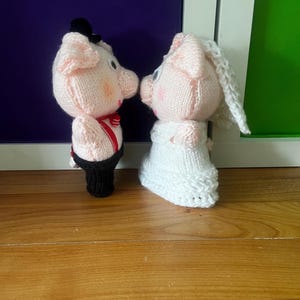 May include: Two knitted pig figurines, one dressed as a groom in a black tuxedo with a red bow tie and braces, and the other as a bride in a white dress and veil. The pigs are facing each other, suggesting a wedding theme. The background is a mix of purple and green.