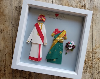 May include: A framed paper art piece featuring a couple's wedding attire. The groom's outfit is white with red and gold accents, and the bride's is green and gold. The frame includes the names "Saksham" and "Aisha" and the date "November 10th 2020".