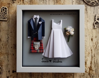 May include: A framed paper art piece depicting a wedding couple. The groom is wearing a blue suit with a kilt and the bride is wearing a white dress. The frame has a gray background with the text "1st Anniversary - 3rd August 2023" and "Jamie ♥ Andrea".
