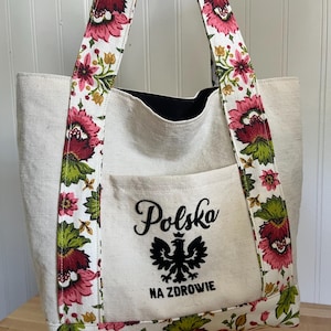 Op de afbeelding: Een crèmekleurige draagtas met bloemmotiefbanden en -bies. De banden en bies hebben een witte achtergrond met roze en rode bloemen en groene bladeren. De tas heeft een voorvak met het woord "Polska" en een zwart adelaarsymbool.