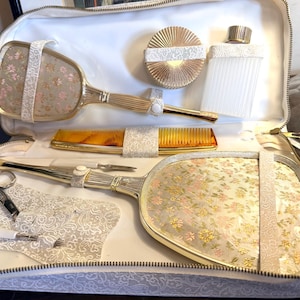 Puede incluir: Un set de tocador en un estuche con cremallera que incluye dos espejos de mano con reverso de tela floral, un peine dorado, un pequeño recipiente redondo con tapa, una botella blanca estriada, tijeras de metal y una herramienta, sujetos por correas.