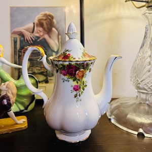 May include: White ceramic teapot with a floral rose pattern in shades of red, pink, and yellow. The teapot has a curved spout, a handle with gold accents, and a matching lid. Decorative figurines and a framed picture are in the background.