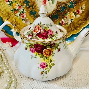 Könnte beinhalten: Weiße Porzellan-Teekanne mit einem Blumendesign aus roten, rosa und gelben Rosen, akzentuiert mit goldenen Verzierungen. Die Teekanne hat einen geschwungenen Griff und einen Deckel mit Knauf. Eine Perlenkette und ein dekoratives Tablett befinden sich im Hintergrund.