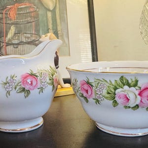 Może przedstawiać: Zestaw dzbanek na mleko i cukiernica z białej porcelany z delikatnym kwiatowym wzorem różowych róż i małych białych kwiatków. Obie części wykończone są cienką złotą obwódką wzdłuż krawędzi i uchwytów, co nadaje im klasyczny wygląd.