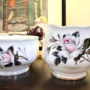 Może przedstawiać: Biały zestaw porcelanowy składający się z cukiernicy i dzbanuszka na śmietankę, ozdobiony delikatnym motywem różowych róż z ciemnozielonymi i czarnymi liśćmi. Oba naczynia mają faliste brzegi oraz złocone podstawy, a dzbanuszek posiada wygięte ucho.