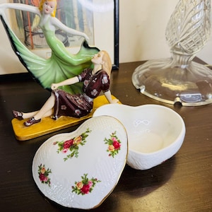 Può includere: Un portagioie bianco a forma di cuore con un coperchio decorato con rose rosse e gialle e foglie verdi. La scatola ha un bordo dorato. Una statuina di porcellana di una donna con un vestito verde e un'altra con un vestito marrone sono sullo sfondo.