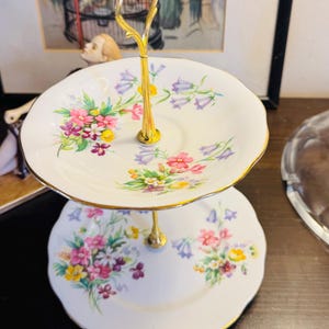 Peut inclure: Un présentoir à deux étages composé d'assiettes en porcelaine blanche aux bords festonnés et d'une poignée en métal doré. Chaque assiette est ornée d'un motif floral coloré de fleurs roses, jaunes et violettes avec des feuilles vertes.