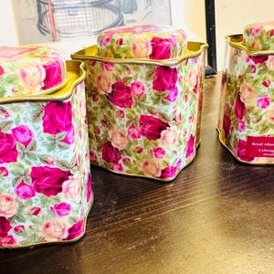 May include: Three square metal canisters with rounded corners and matching lids, featuring a vibrant floral pattern of pink and yellow roses on a light background. The canisters have gold-toned rims and a label on the rightmost one reading Royal Albert.