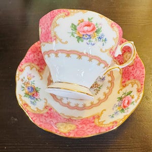 May include: A white porcelain teacup and saucer set featuring a vibrant pink floral pattern with gold trim. The cup and saucer are decorated with detailed bouquets of pink roses and small blue flowers, accented by ornate gold scrollwork borders.