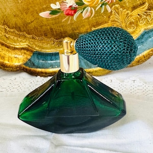 Pode incluir: Um frasco de perfume vintage com um corpo de vidro verde esmeralda facetado. Possui um atomizador dourado e uma pêra tecida de cor verde-azulado. O frasco está sobre um fundo de uma bandeja com acabamentos dourados e detalhes florais.