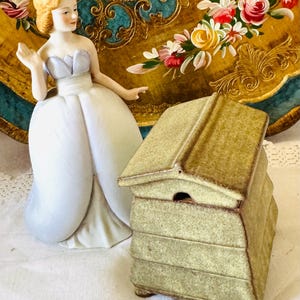 May include: A porcelain figurine of a person with blonde hair wearing a light blue and white gown, positioned next to a small, textured, light green ceramic beehive-shaped container with a removable lid and a small opening at the front.