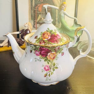 以下が含まれることがあります： ピンク、黄色、赤の色合いのバラの花柄が施された白いセラミックティーポット。 ティーポットは金色のトリムと湾曲したハンドルが付いています。 蓋には白いつまみが付いています。 クラシックなティーパーティーアイテムです。