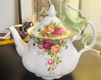 May include: White ceramic teapot with a floral rose design in shades of pink, yellow, and red. The teapot has a gold trim and a curved handle. The lid is topped with a white finial. A classic tea party item.