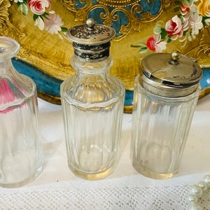 Può includere: Tre bottiglie di vetro trasparente con tappi argentati. Le bottiglie hanno un design a coste e sono appoggiate su una superficie bianca.