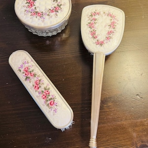 May include: A vintage vanity set comprising a round lidded container, a hand mirror, and a brush. Each item has a cream surface adorned with a floral pattern of pink roses and purple details. The handles and rims are silver-toned.
