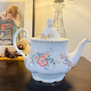 Könnte beinhalten: Eine weiße Porzellanteekanne mit gewelltem Körper, passendem Deckel mit spitzem Knauf und geschwungenem Henkel. Die Teekanne ist mit einem bunten Blumenmuster aus rosa, blauen und gelben Blüten verziert, das an den Rändern mit Gold verziert ist.