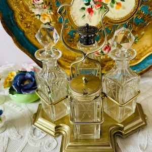 May include: An antique vanity set with a brass base and frame, holding four clear glass bottles and a small jar. The bottles have faceted crystal stoppers. The set is displayed on a white lace surface, with a floral tray in the background.