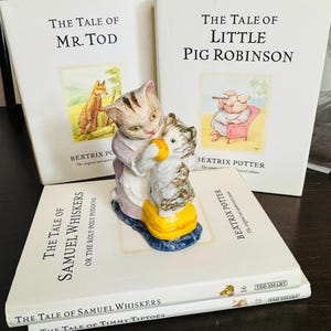 Puede incluir: Una colección de libros de Beatrix Potter, incluyendo "The Tale of Mr. Tod" y "The Tale of Little Pig Robinson". Una figura de cerámica de dos gatos está encima de los libros. Los libros tienen cubiertas blancas con ilustraciones y texto.