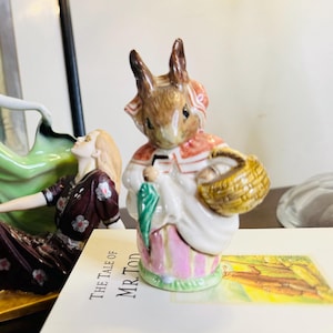 May include: A ceramic figurine of a rabbit wearing a red and white outfit, holding a basket and a green object. The rabbit has brown ears and is standing on a base with pink stripes. The figurine is placed on a book titled "The Tale of Mr. Tod".