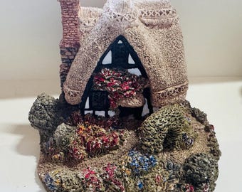 Może przedstawiać: Miniaturowa figurka wiejskiego domku z dachem z trzciny, z ceglanym kominem, otoczona zielonymi krzewami i kolorowymi kwiatami. Domek ma czarne drzwi i okna oraz białą obwódkę wokół dachu.
