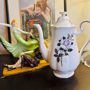 May include: A white porcelain coffee pot with a floral design featuring a pink rose and gray leaves. The pot has a curved handle and a gold trim. Decorative figurines are in the background.