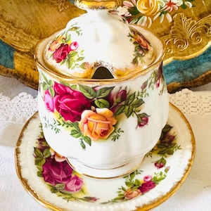 Könnte beinhalten: Eine weiße Zuckerdose aus Keramik mit Deckel und passender Untertasse, verziert mit einem floralen Rosenmuster in Rot-, Rosa- und Gelbtönen. Die Ränder sind goldfarben verziert. Die Schüssel hat eine kleine Öffnung für einen Löffel.
