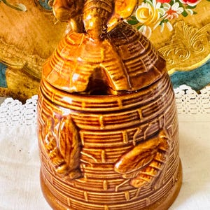 Puede incluir: Tarro de miel de cerámica marrón con tapa, con forma de colmena. El tarro presenta diseños en relieve de abejas y hojas. La tapa está coronada con una figura de abeja. El tarro está sobre una superficie blanca.