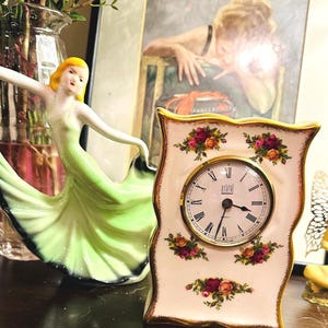 Puede incluir: Un reloj decorativo de porcelana crema con borde dorado y esfera con números romanos. El reloj está decorado con motivos florales. También se ve una figura de cerámica de una mujer con un vestido verde.