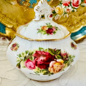 May include: A white porcelain sugar bowl with a lid, decorated with a rose floral pattern in red, pink, and yellow. The rim and handle are trimmed with gold. The bowl is on a white lace surface.