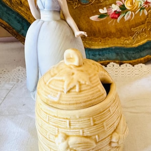 May include: A light yellow ceramic honey pot with a lid, featuring a basket weave design and a bee motif. The pot sits on a white surface. A porcelain figurine and a decorative gold-framed plate are in the background.