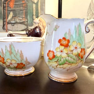 Puede incluir: Un juego de lechera y azucarero de porcelana blanca con un diseño floral pintado a mano con flores naranjas y amarillas, follaje verde y bordes dorados en las bases y aberturas. Ambas piezas presentan una forma redondeada.
