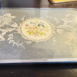 May include: A rectangular serving tray with a chrome metal frame and handles. The centre features a glass panel with a white lace floral overlay and a circular fabric insert displaying a colourful embroidered bouquet of pink, blue, and yellow flowers.