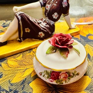 Puede incluir: Una caja de baratijas de cerámica blanca con una rosa roja en la tapa y diseños florales. La caja tiene un borde dorado y está sobre una superficie con estampado floral colorido. Una figura está en el fondo.