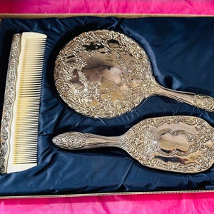 May include: A vintage vanity set comprising a silver-toned hand mirror, hairbrush, and comb. The set features ornate floral detailing and is presented in a box with a blue satin lining. The comb has a cream-coloured handle.