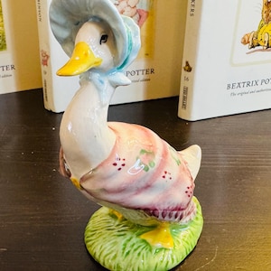 May include: A ceramic figurine of a duck wearing a blue bonnet and a pink dress with floral accents. The duck stands on a green grassy base. The figurine is a collectible item.