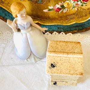 May include: A porcelain figurine of a person with light brown hair wearing a pale blue dress, positioned next to a square, speckled ceramic box featuring small black bee illustrations on the side and front panels of the lid and base.