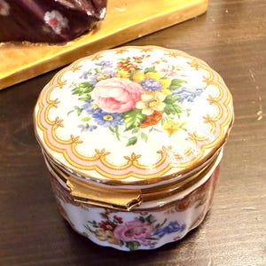Może przedstawiać: Okrągłe pudełko na drobiazgi z porcelany z falistym brzegiem i zawiasowym zamknięciem z metalu w kolorze złota. Pokrywka i podstawa mają białe tło ozdobione kolorowymi bukietami kwiatów, w tym różowymi różami, żółtymi żonkilami i niebieskimi kwiatami.