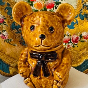 May include: A ceramic teddy bear cookie jar with a glossy, brown finish and a black bow tie. The bear has large, round ears and a sweet expression. It sits on a white cloth with embroidered flowers, against a floral background.