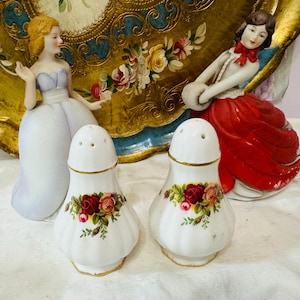 May include: Two white porcelain salt and pepper shakers with a floral rose design. The shakers have a gold trim and a classic shape. Decorative figurines and a gold-trimmed tray are in the background.