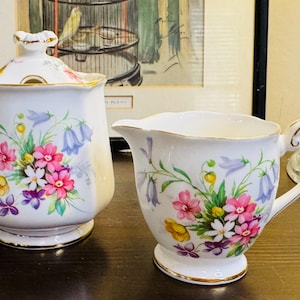 May include: A white porcelain sugar bowl with a lid and a creamer, decorated with a floral design featuring pink, purple, yellow, and blue flowers. The rims and bases are trimmed with gold. The creamer has a curved handle.