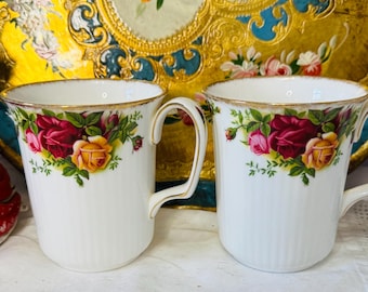 May include: Two white ceramic mugs with gold trim and floral designs featuring red and yellow roses. The mugs have a ribbed texture and curved handles. Decorative figurines and a gold tray with floral details are in the background.