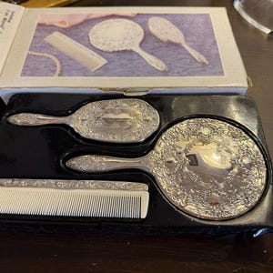 Peut inclure: Un ensemble de toilette de trois pièces dans un plateau noir moulé, comprenant un miroir à main argenté, une brosse à cheveux et un peigne avec une base en plastique blanc. L'ensemble est présenté dans une boîte en carton illustrée.