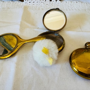 May include: A gold-toned metal hand mirror, a round open compact case with an interior mirror, a fluffy white powder puff with a small yellow ribbon, and a closed gold-toned metal round compact case, all arranged on a white fabric surface.