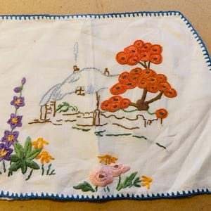 Könnte beinhalten: Ein weißes besticktes Geschirrtuch mit blauem Rand. Das Design zeigt ein kleines Haus, einen Baum mit orangefarbenen Blättern und lila und gelben Blumen.
