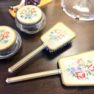 Op de afbeelding: Een kapset bestaande uit twee ronde glazen potten met bloemendeksels, een rechthoekige haarborstel en een rechthoekige handspiegel. Elk item heeft een crèmekleurig paneel met een geschilderd ontwerp van roze rozen en blauwe bloemen.