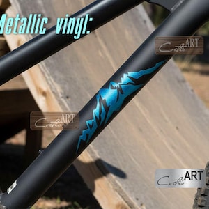 Peut inclure: Cadre de vélo noir avec un autocollant en vinyle métallique bleu en forme de montagne. Le texte "Metallic vinyl" est en bleu clair en haut. L'image comprend également le texte "Cortis ART" sur une plaque en bois et une plaque argentée.