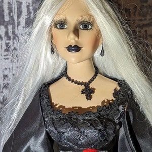 Puede incluir: Una muñeca de porcelana con el pelo largo y blanco, vestida con un vestido negro con ribete de encaje y una cruz roja. Lleva maquillaje oscuro, pendientes negros y un collar de cuentas negras con un colgante de cruz.